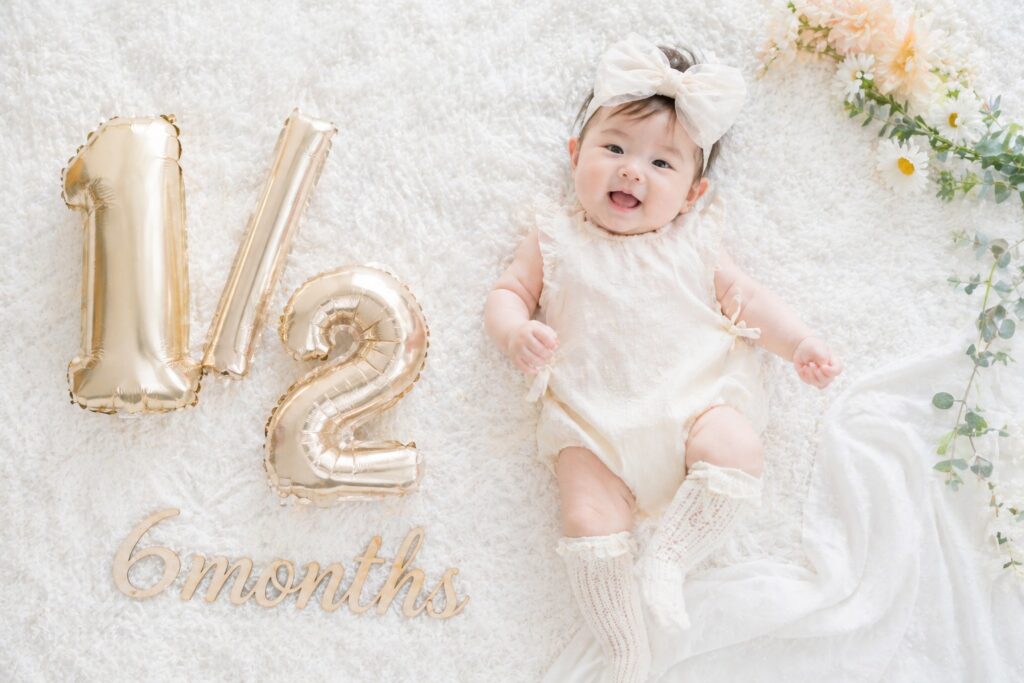 白いラグの上にリボンをつけた赤ちゃん、数字バルーンと「6months」レターバナーが並んだ俯瞰写真