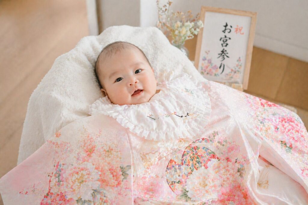 明るい部屋の白い床の上で、お宮参りの掛け着（祝い着）をまとった赤ちゃんを抱っこしているシーン